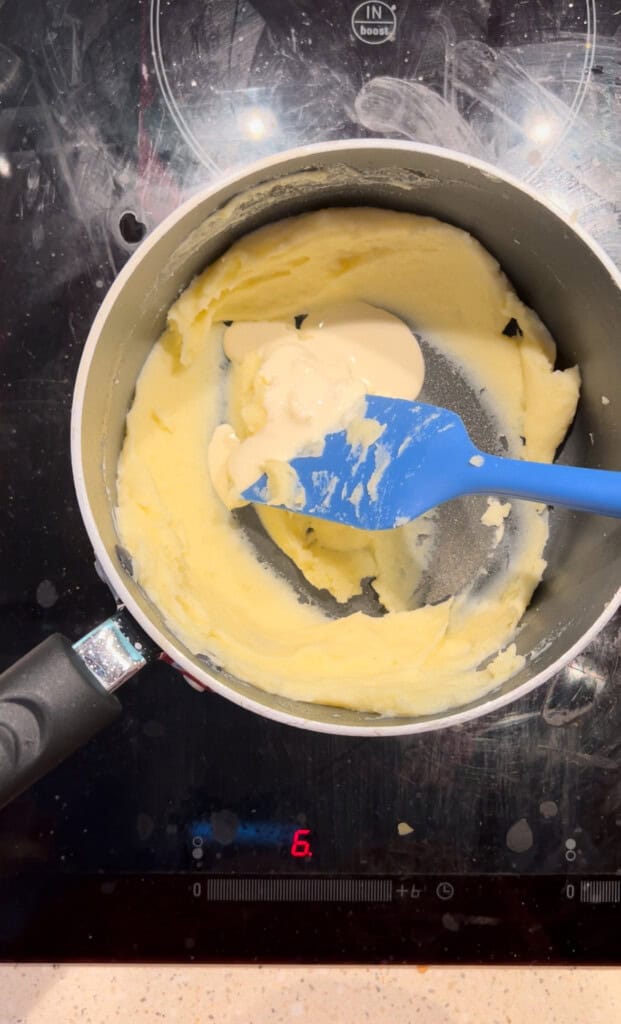 A saucepan with creamy, partially mixed batter and a blue spatula sits on a black hob set to level 6. Some light-coloured sauce or cream is dolloped on top of the mixture.