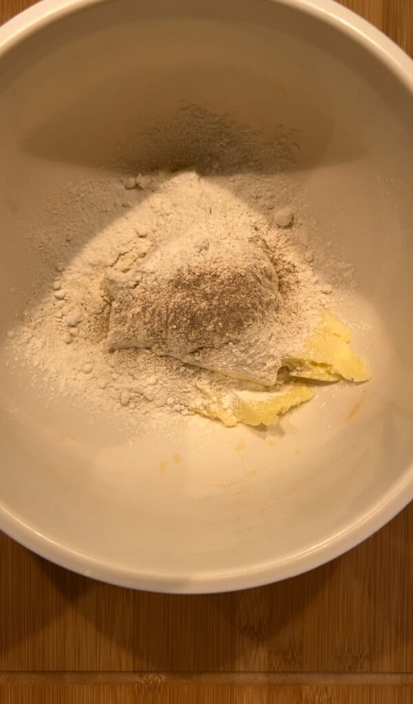 A white mixing bowl containing flour, black pepper, and softened butter, all unmixed and ready for preparation, sits on a wooden surface.