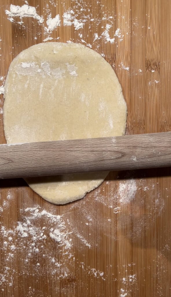 A wooden rolling pin rolling out pastry on a floured wooden surface. Icing sugar is scattered around the pastry.