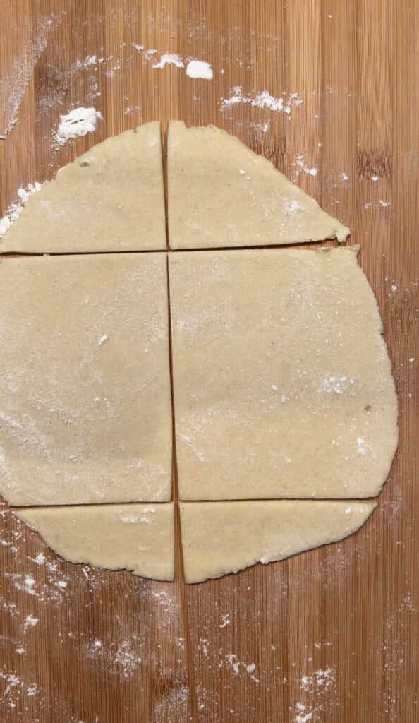 A round sheet of dough on a wooden surface is lightly dusted with flour and cut into six square pieces.
