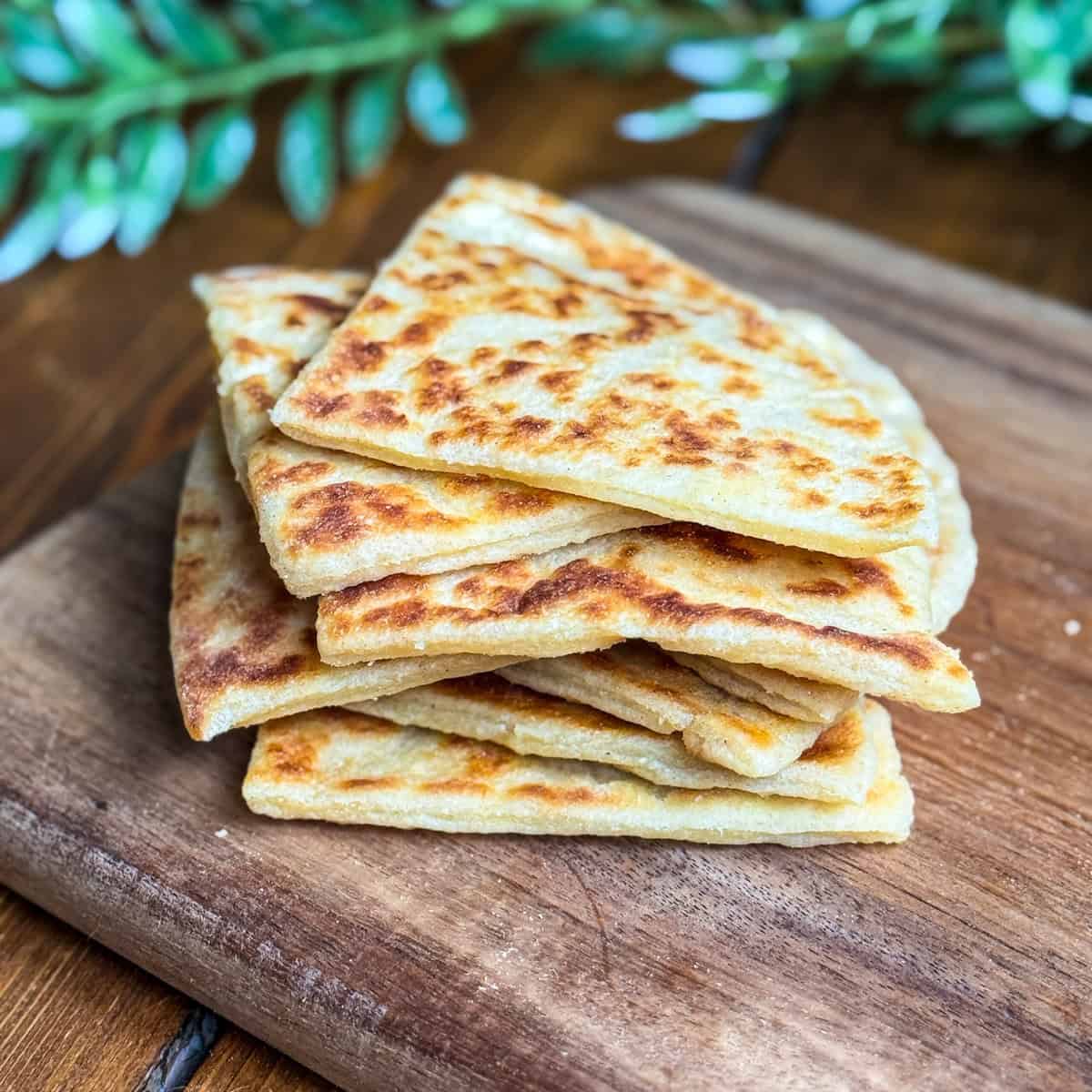 A stack of golden-brown, triangular flatbreads sits on a wooden cutting board, with a leafy green plant blurred in the background.