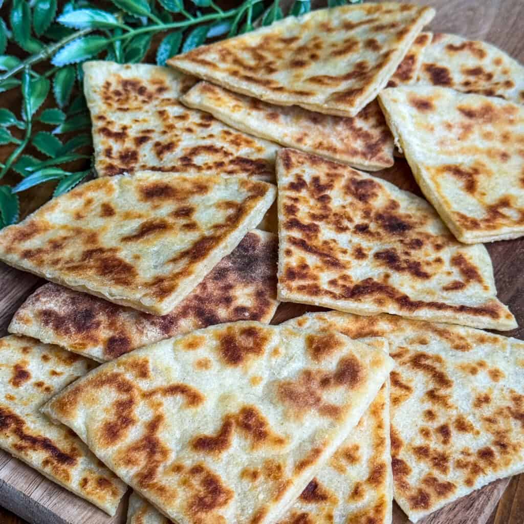 Golden-brown, pan-fried potato scones cut into triangles are arranged on a wooden board, with green leaves in the background.