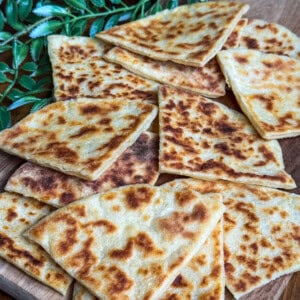 Golden-brown, pan-fried potato scones cut into triangles are arranged on a wooden board, with green leaves in the background.