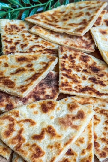 Golden-brown, pan-fried potato scones cut into triangles are arranged on a wooden board, with green leaves in the background.