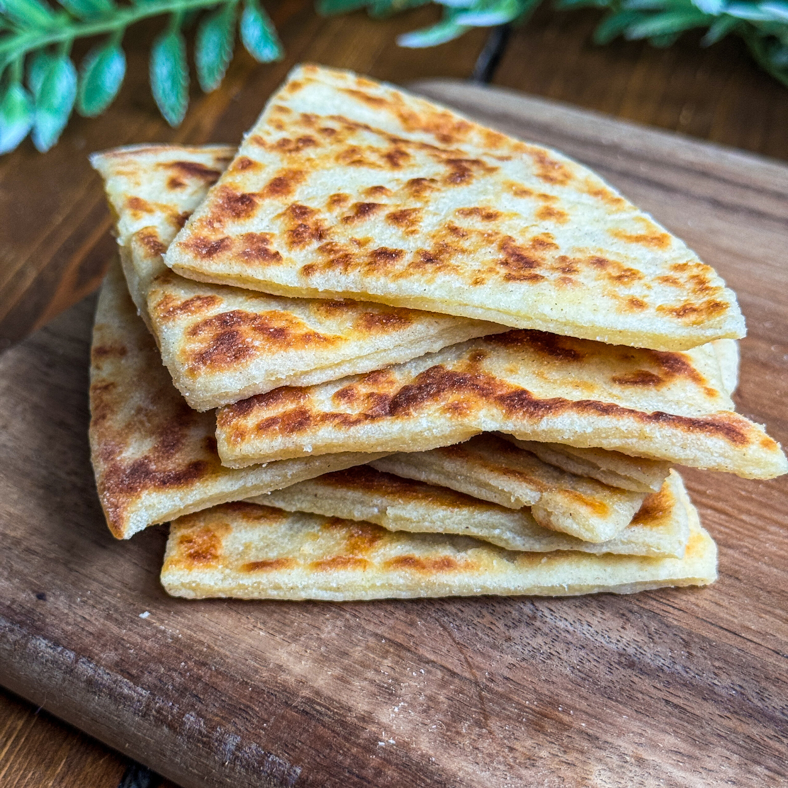 A stack of golden-brown potato scones sits on a wooden board with green leafy plants in the background.