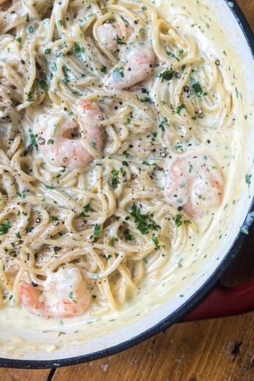 A creamy pasta dish with spaghetti and shrimp in a light-colored sauce, garnished with freshly chopped parsley and black pepper, served in a large, round red-handled pan on a wooden table.