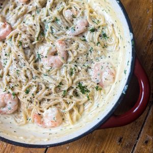 A creamy pasta dish with spaghetti and shrimp in a light-colored sauce, garnished with freshly chopped parsley and black pepper, served in a large, round red-handled pan on a wooden table.