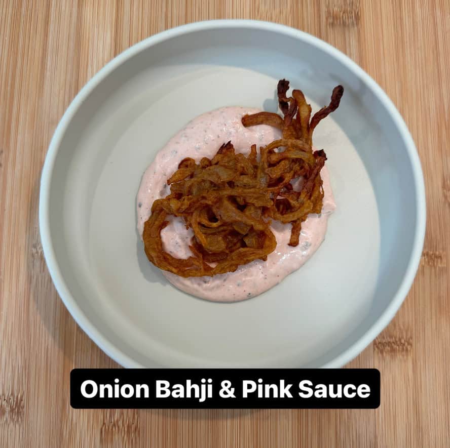 A white bowl contains crispy onion bhajis on a serving of pink sauce. The bowl is placed on a wooden surface. Text at the bottom reads, Onion Bahji & Pink Sauce.