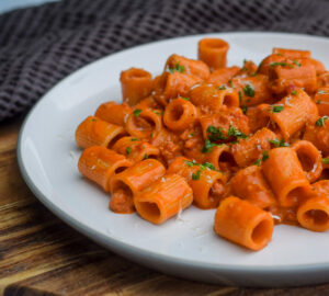 A white plate filled with rigatoni pasta covered in a creamy tomato sauce, garnished with chopped herbs. The plate is on a wooden surface, with a dark textured fabric in the background.