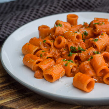 A white plate filled with rigatoni pasta covered in a creamy tomato sauce, garnished with chopped herbs. The plate is on a wooden surface, with a dark textured fabric in the background.
