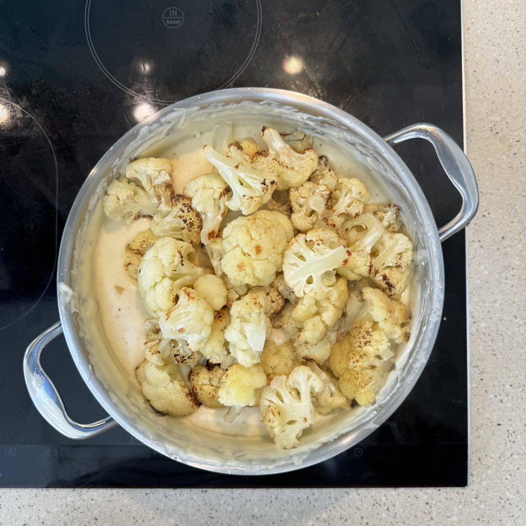 A pot filled with roasted cauliflower florets in a creamy white sauce. The cauliflower is lightly browned, indicating it has been cooked. The pot has metal handles and is placed on a dark stovetop.