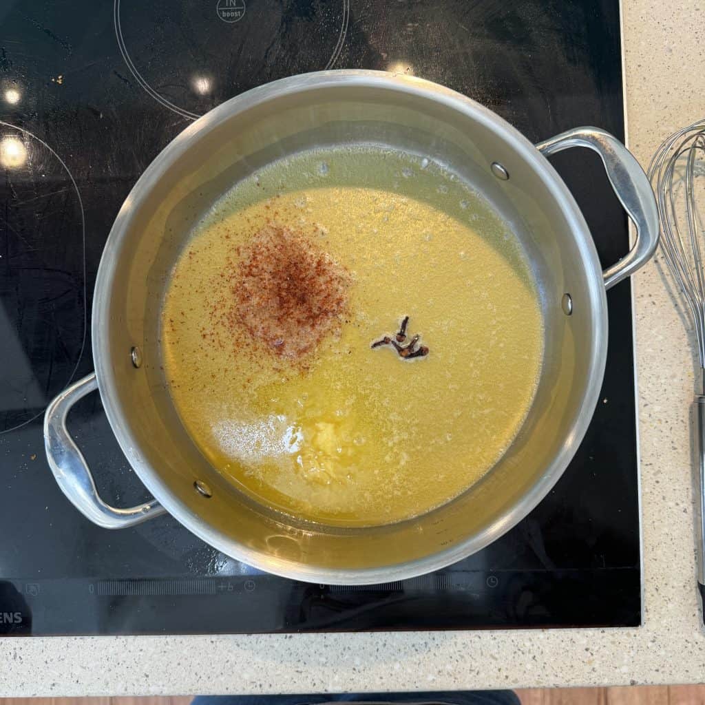 A pot on a stove contains a yellow liquid, possibly broth or soup, with spices including a sprinkle of red powder and a few dark whole spices. A whisk is visible on the countertop beside the pot.