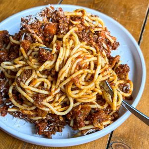 A plate of spaghetti Bolognese topped with grated cheese sits on a wooden table. The pasta is mixed with a rich, chunky tomato and meat sauce. A fork rests on the edge of the white plate.