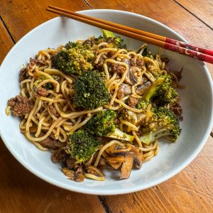 A bowl of stir-fried noodles with beef, broccoli, and mushrooms is garnished with red chopsticks placed on the rim. The dish is served in a white bowl on a wooden table.