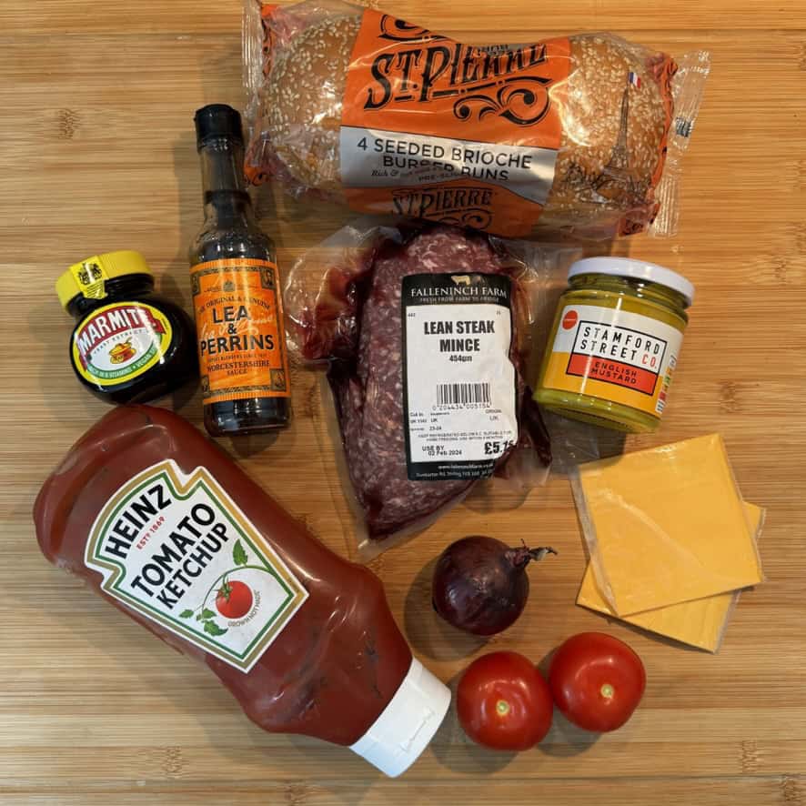 A wooden surface displays burger ingredients: four seeded brioche buns, a pack of lean steak mince, a bottle of Worcestershire sauce, Marmite, pickles, ketchup, processed cheese slices, two tomatoes, and a red onion.