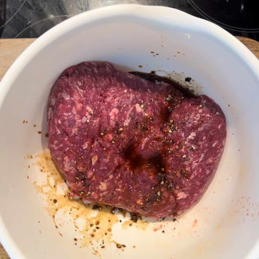 A white bowl containing raw ground beef, seasoned with black pepper and sauce. The beef is reddish-brown and sits on a kitchen countertop.