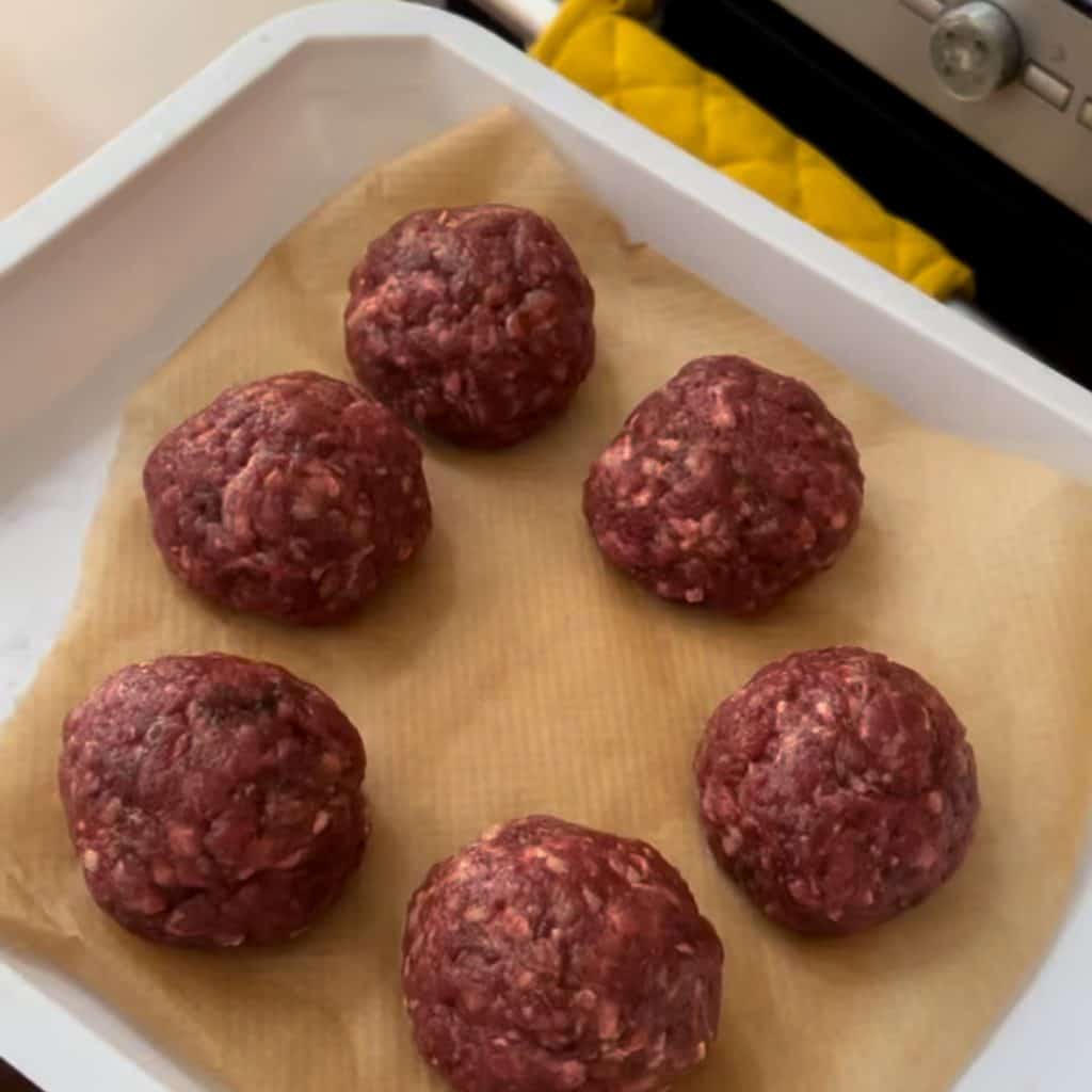 Six raw ground beef balls sit on parchment paper in a white baking tray, placed on a kitchen counter near an oven. A yellow oven mitt is visible beside the tray.