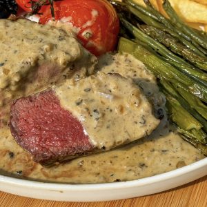 Close-up of a steak topped with creamy peppercorn sauce, served alongside whole grilled tomatoes, asparagus, and a portion of fries, all on a white plate. The steak is cooked medium, revealing a pink center.