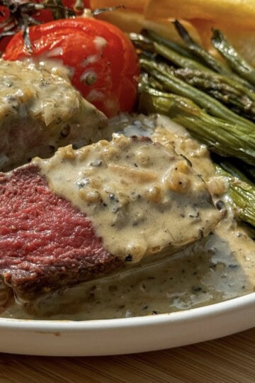 A plate featuring a juicy steak covered in creamy peppercorn sauce, accompanied by roasted cherry tomatoes, green beans, and crispy potato chips. The steak is cooked medium-rare, revealing a pink center.