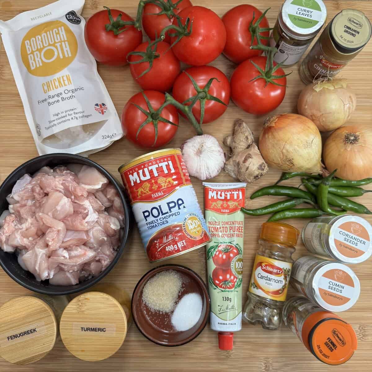 A flat lay of ingredients for a curry, including fresh tomatoes, onions, green chilies, garlic, ginger, chicken pieces, broth, canned and tubed tomato, spices in jars and bowls, and various spice containers with labels.