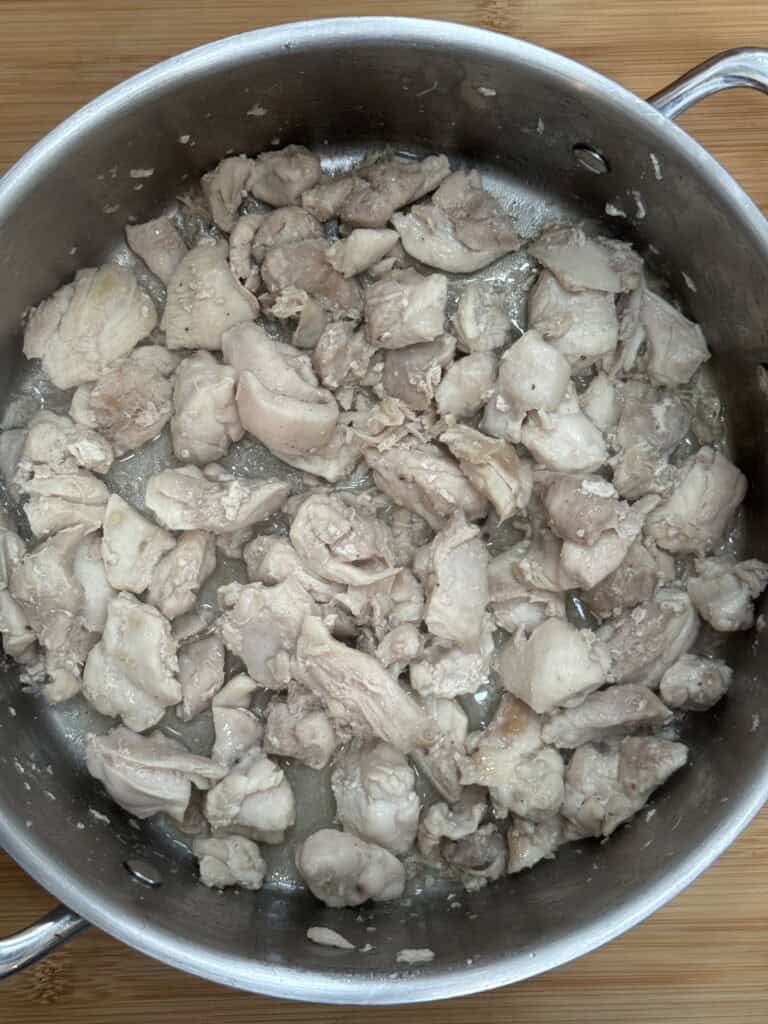 Chopped pieces of cooked chicken simmering in a stainless steel pot, with visible juices collecting at the bottom, on a wooden surface.