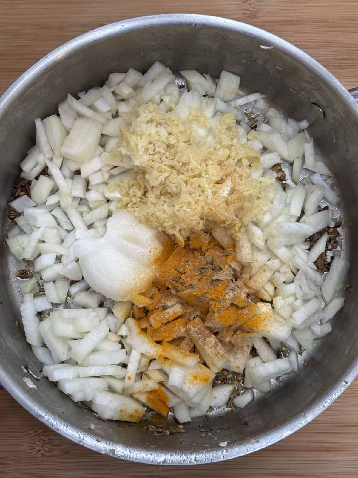 A metal pot contains chopped onions, grated ginger, ground turmeric, ground cinnamon, salt, and other spices on a wooden surface, ready to be mixed and cooked.