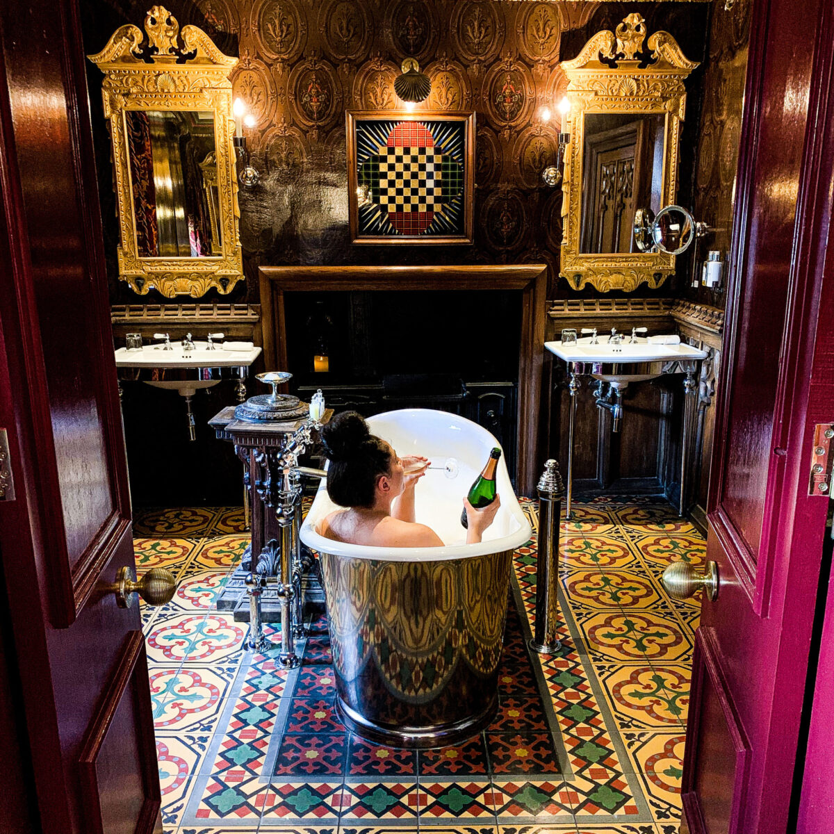 A person is relaxing in a luxurious bathtub with a bottle of wine in hand, surrounded by ornate decor. The room features vintage-style patterned tiles, two elegant mirrors, sinks, and warm lighting.