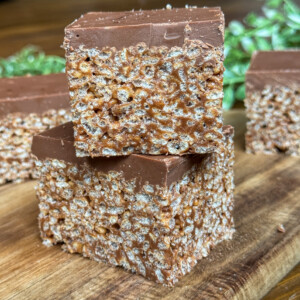 Two chocolate-topped crispy rice treats are stacked on a wooden surface, with more treats in the background and some green leaves out of focus.