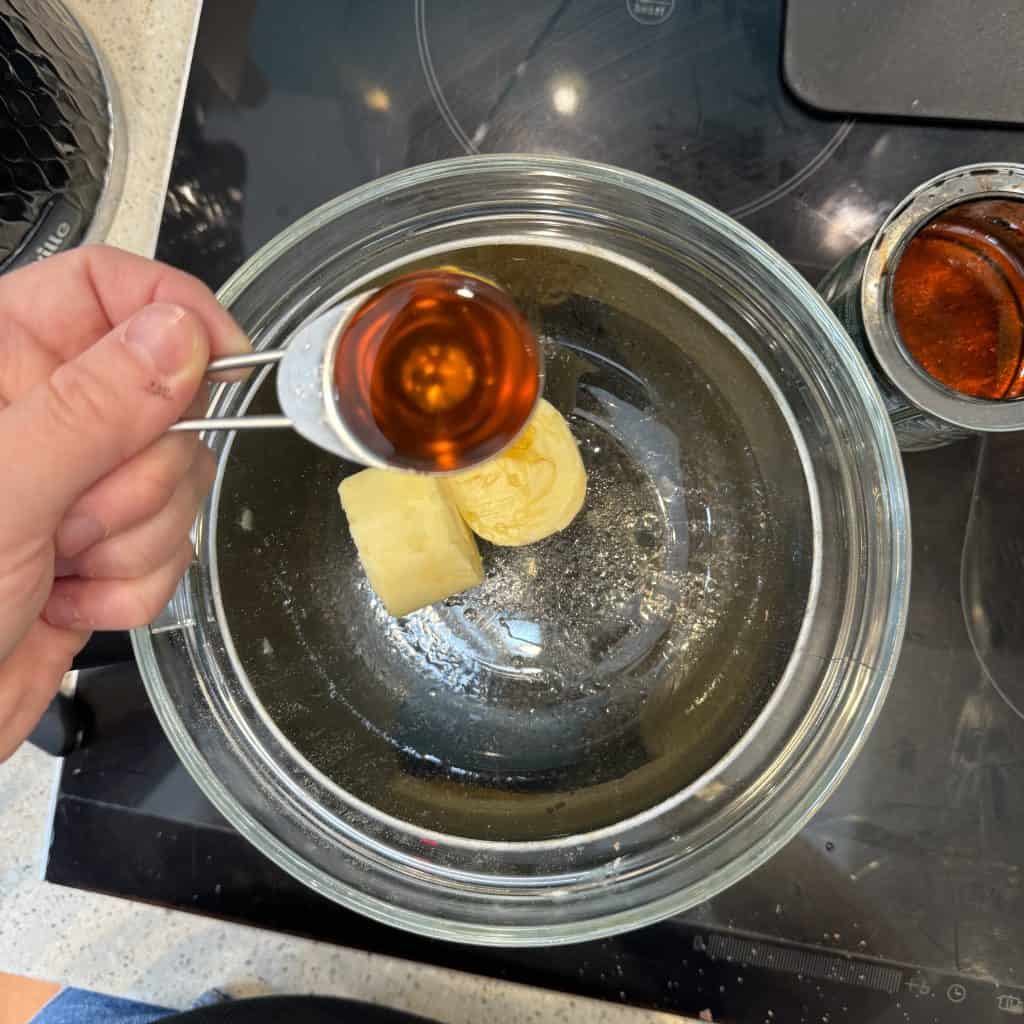 A person is adding a tablespoon of brown liquid, possibly vanilla extract, to a glass bowl containing melted butter and sugar. The bowl is on a black stovetop. Another small jar with brown liquid is on the side.