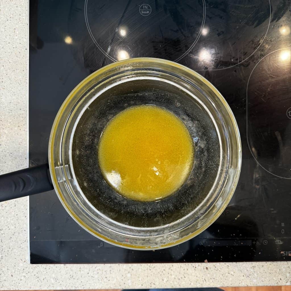 A saucepan on an electric stovetop contains melted butter, creating a circular yellow pool in the center of the clear liquid. The black stovetop surface contrasts with the countertops light, speckled pattern.