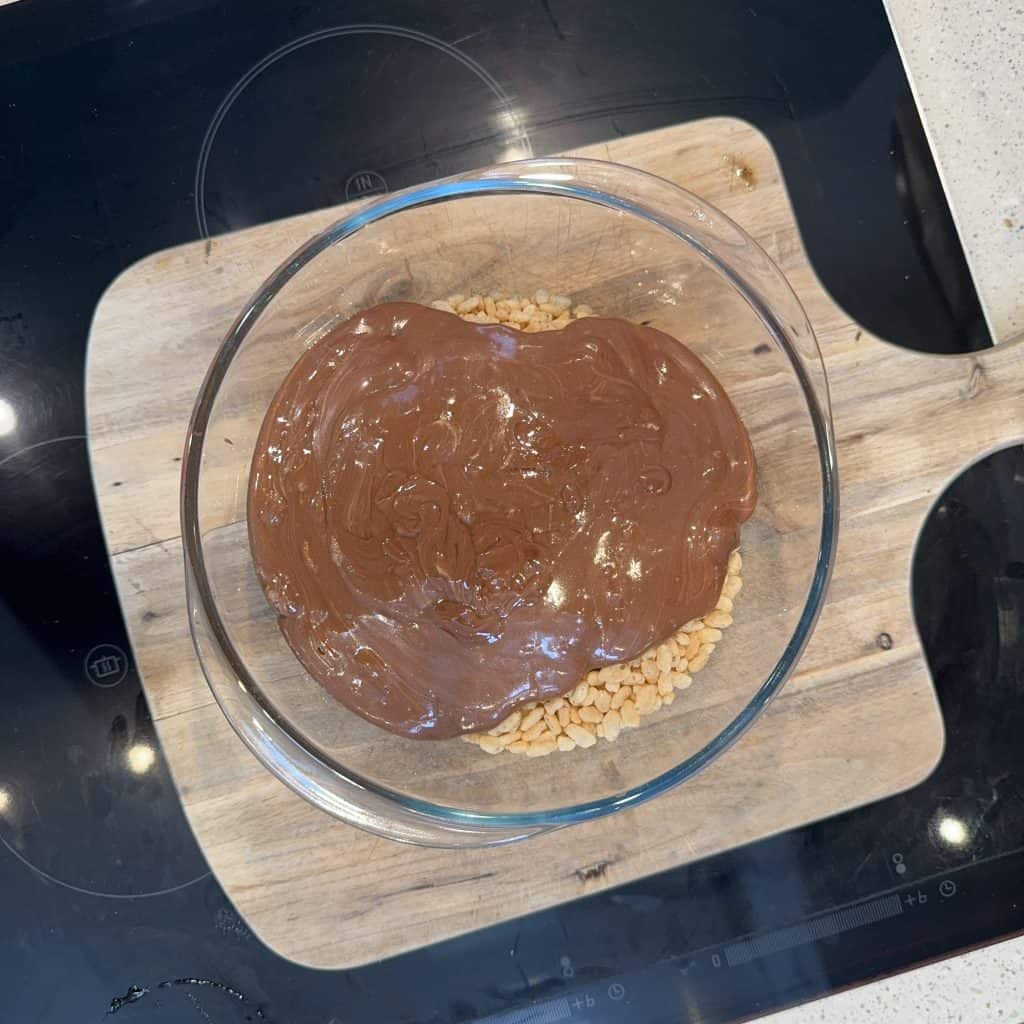 A glass bowl on a wooden board contains a layer of chocolate spread over a base of rice cereal. The bowl is placed on a black induction stovetop.
