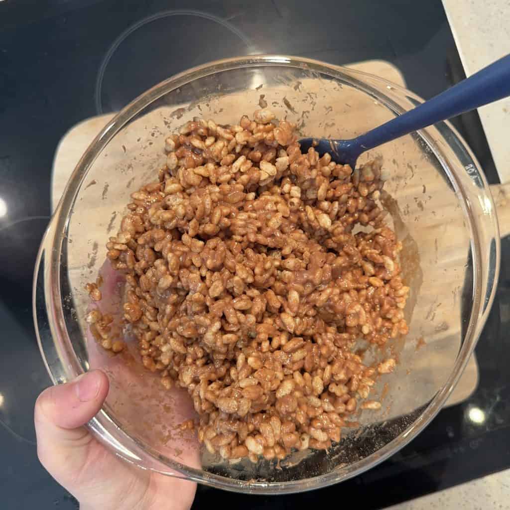 A glass bowl filled with a mixture of puffed rice cereal and chocolate, being stirred with a blue spatula. The bowl is held over a black stovetop.