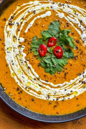 A bowl of creamy orange soup garnished with a swirl of cream, cilantro leaves, and slices of red chili peppers. The soup is accented with black pepper and a drizzle of olive oil, placed on a wooden surface.