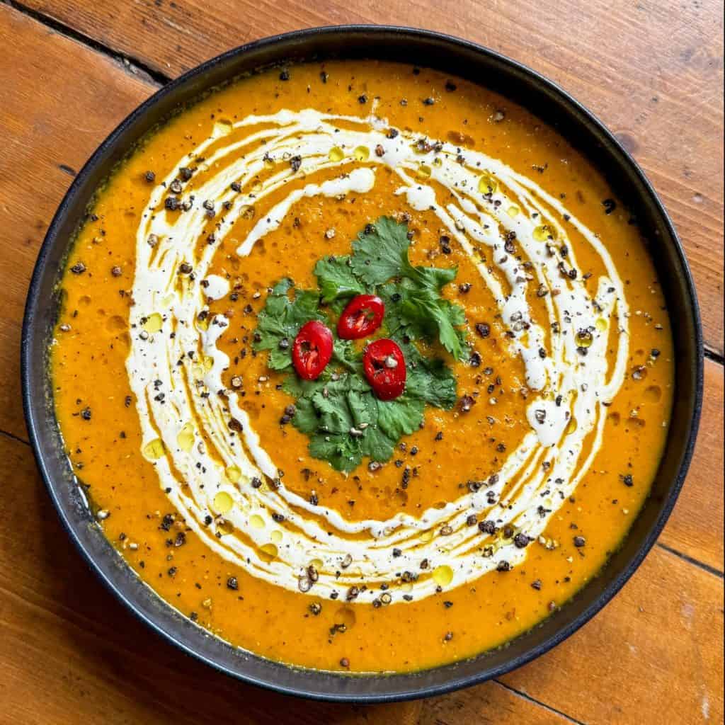 A bowl of creamy orange soup garnished with a swirl of white cream, fresh cilantro leaves, black pepper, and red chili slices, set on a wooden surface.