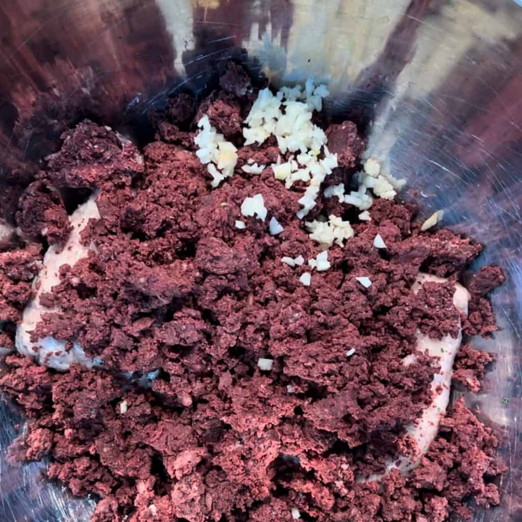 Close-up of a metal mixing bowl containing crumbled chunks of a dark red mixture and small pieces of minced garlic. The textures and colors contrast against each other, with the garlic scattered on the surface.