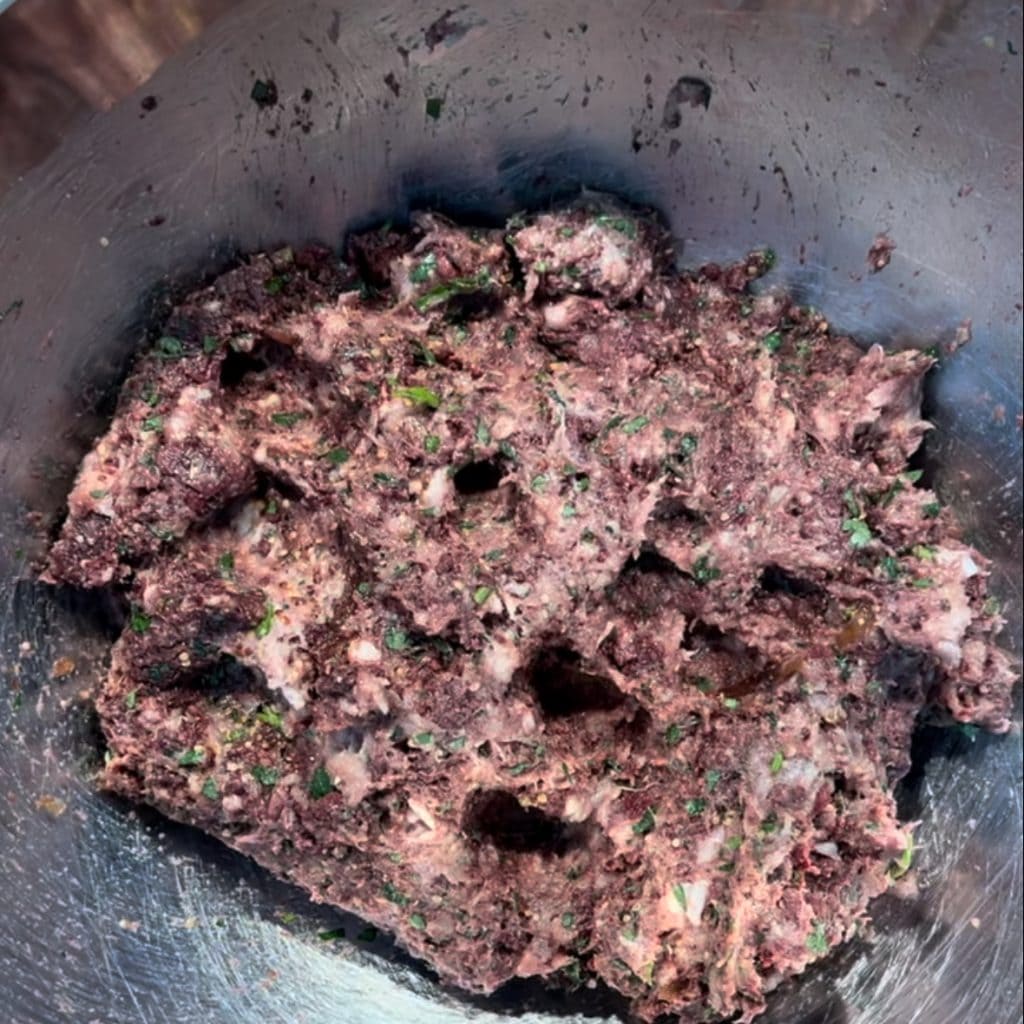 Close-up of a metal bowl containing a mixture of ground meat and herbs. The mixture has a coarse texture with visible pieces of herbs, giving it a speckled appearance. The bowl is slightly reflective, capturing ambient light.
