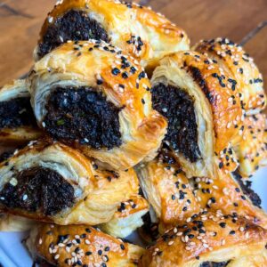 Close-up of a pile of golden-brown, flaky pastry rolls filled with a dark filling, sprinkled with black and white sesame seeds, resting on a white plate. The surface beneath appears to be wooden.