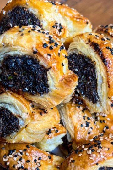 Close-up of a pile of golden-brown, flaky pastry rolls filled with a dark filling, sprinkled with black and white sesame seeds, resting on a white plate. The surface beneath appears to be wooden.