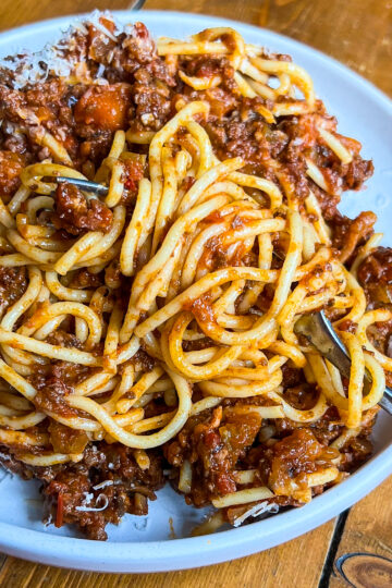 A plate of spaghetti topped with a rich meat sauce, garnished with grated cheese. A fork twirls some pasta. The dish is served on a white plate set on a wooden table.