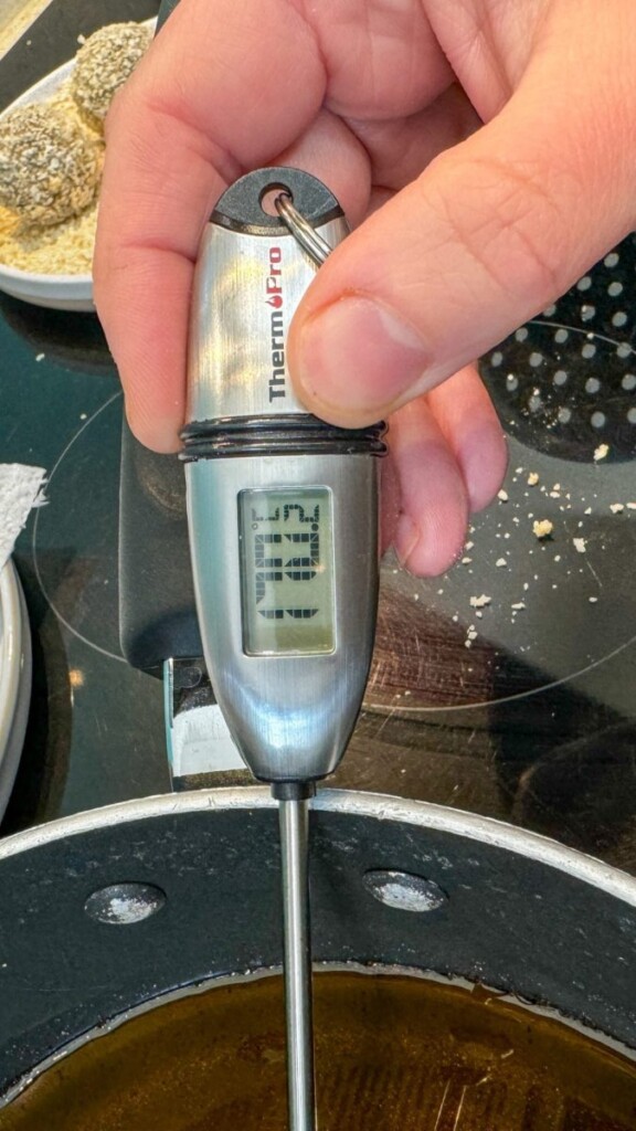 Hand holding a ThermoPro thermometer displaying 110.3°F. The thermometer probe is inserted in a frying pan containing oil. Breadcrumb-covered food items are visible in the background.