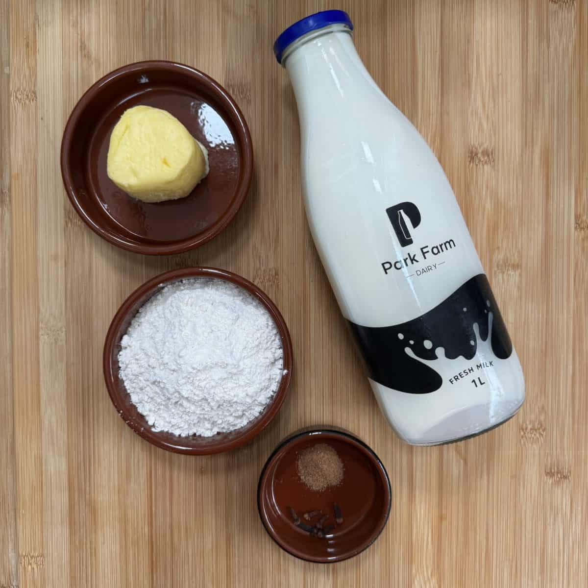 A bottle of fresh milk labeled Park Farm Dairy is on a wooden surface, surrounded by ingredient bowls: a pat of butter, a bowl of flour, and a small dish with ground nutmeg and whole cloves.