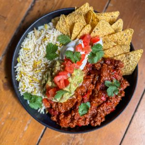A bowl of chili con carne, rice, and tortilla chips topped with diced tomatoes, guacamole, sour cream, and cilantro, placed on a wooden surface.