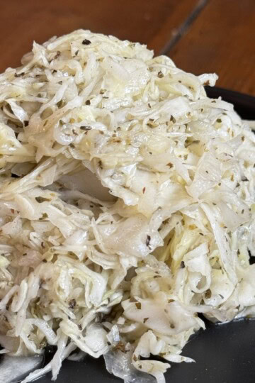 A mound of sauerkraut seasoned with herbs and spices on a black plate. The shredded cabbage appears moist and slightly glistening, with bits of spices visible throughout. The plate sits on a wooden surface.
