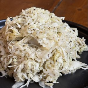 A mound of sauerkraut seasoned with herbs and spices on a black plate. The shredded cabbage appears moist and slightly glistening, with bits of spices visible throughout. The plate sits on a wooden surface.