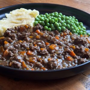 A black plate with minced meat stew containing diced carrots and onions, accompanied by mashed potatoes dusted with breadcrumbs, and a serving of green peas, set on a wooden table.