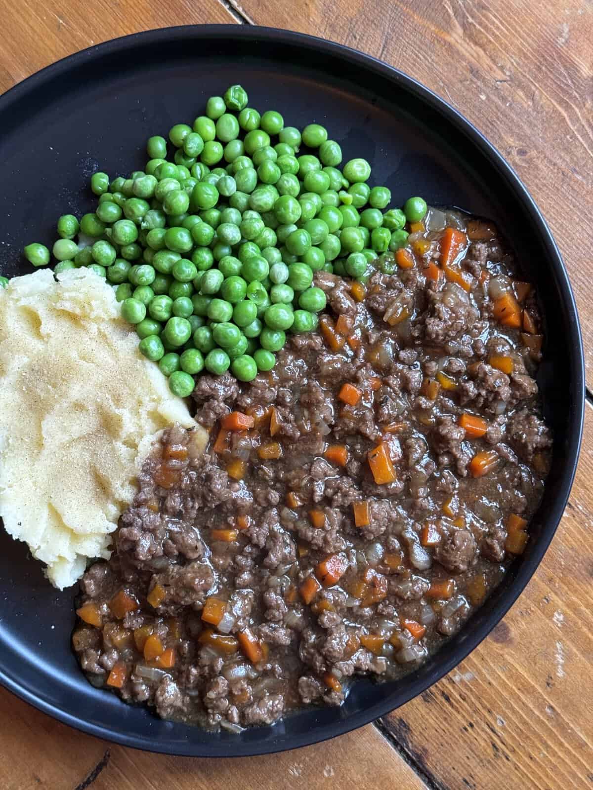 A black plate with mince and tatties: mashed potatoes, green peas, and minced beef with carrots in gravy on a wooden table.