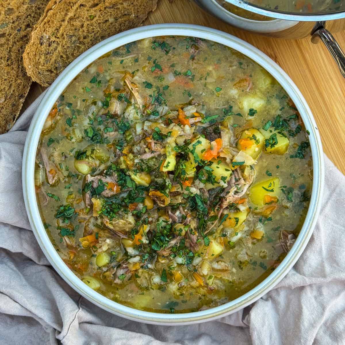 A bowl of hearty scotch broth garnished with chopped herbs sits on a wooden surface, surrounded by slices of rustic bread and a pot in the background.