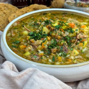 A bowl of hearty scotch broth, garnished with chopped fresh herbs, sits on a light cloth with slices of bread in the background. The soup contains chunks of potato, carrot, and meat.