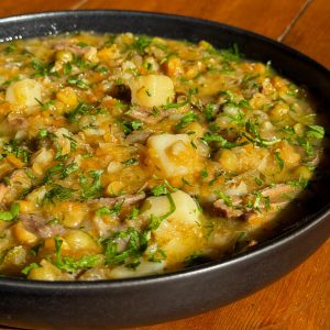 A black bowl filled with a hearty stew, containing chunks of potatoes, chickpeas, shredded meat, and carrots. The dish is garnished with chopped fresh herbs and is placed on a wooden table.