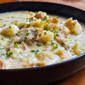 A close-up of a creamy, hearty soup served in a black bowl. The soup contains chunks of potatoes and chicken, garnished with finely chopped green herbs, possibly chives. The texture appears thick and comforting.